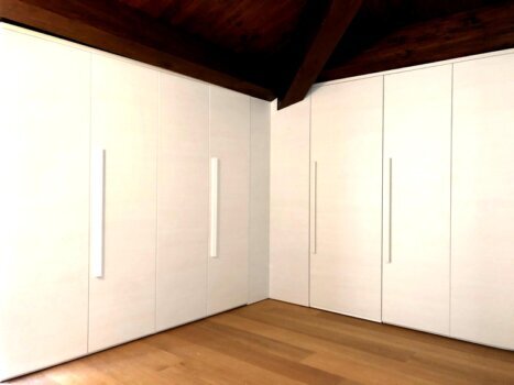 Armadio su misura bianco integrato in una camera mansardata con travi in legno a vista, perfetto per ottimizzare gli spazi con eleganza.