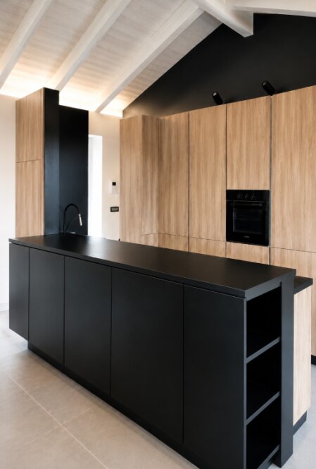Cucina moderna su misura con isola nera, colonne effetto legno, forno integrato e soffitto mansardato con travi bianche.